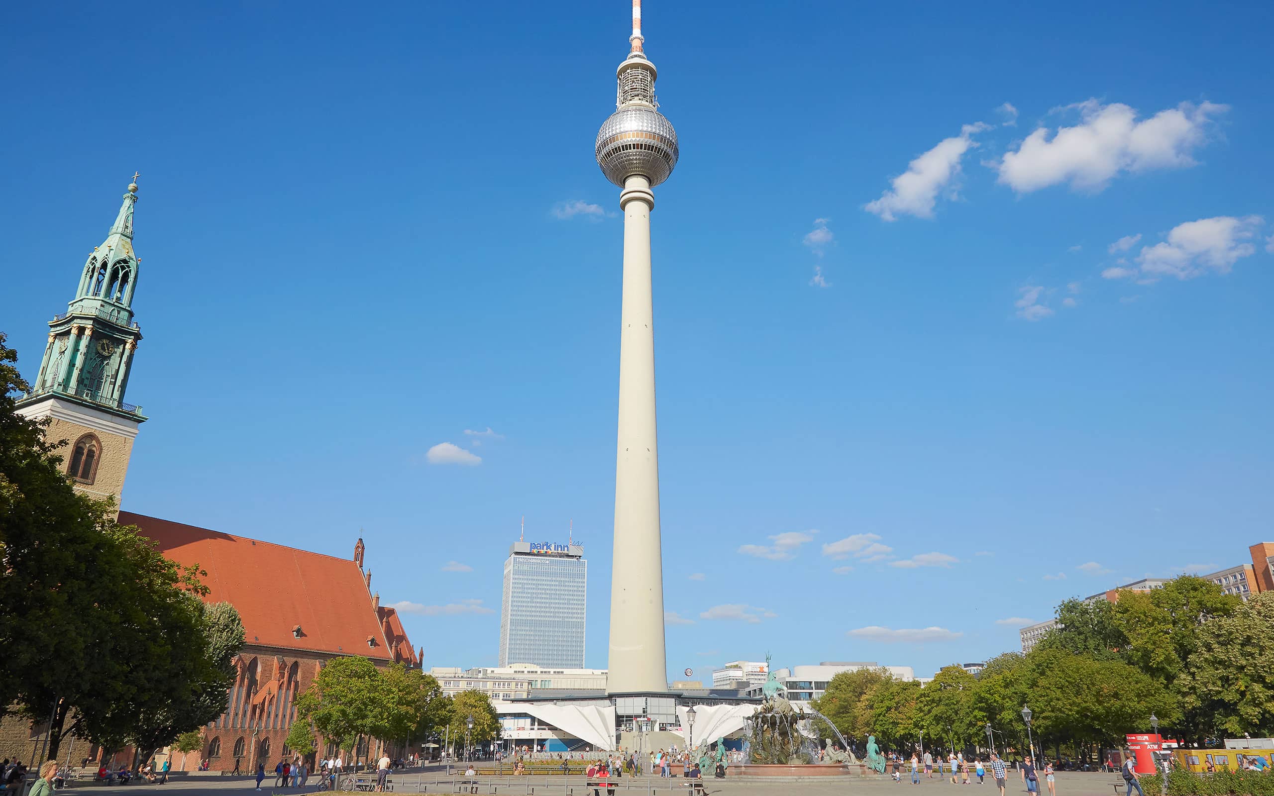 Blick auf den Berliner Fernsehturm am Alexanderplatz bei blauem Himmel, mit Brunnen, Bäumen und umliegenden Gebäuden im Vordergrund.