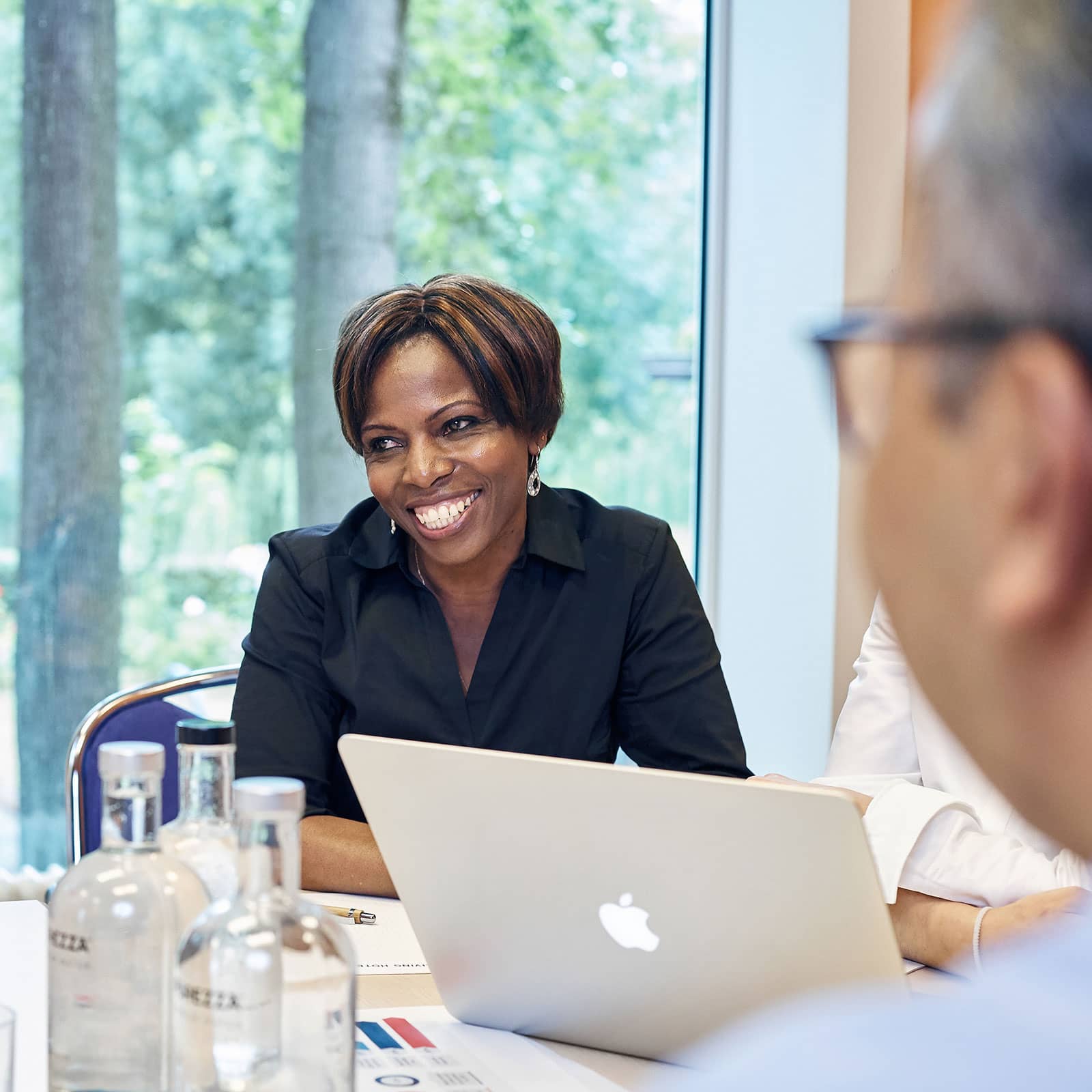 Frau sitzt lächelnd an einem Besprechungstisch und arbeitet an einem Laptop während eines Meetings.
